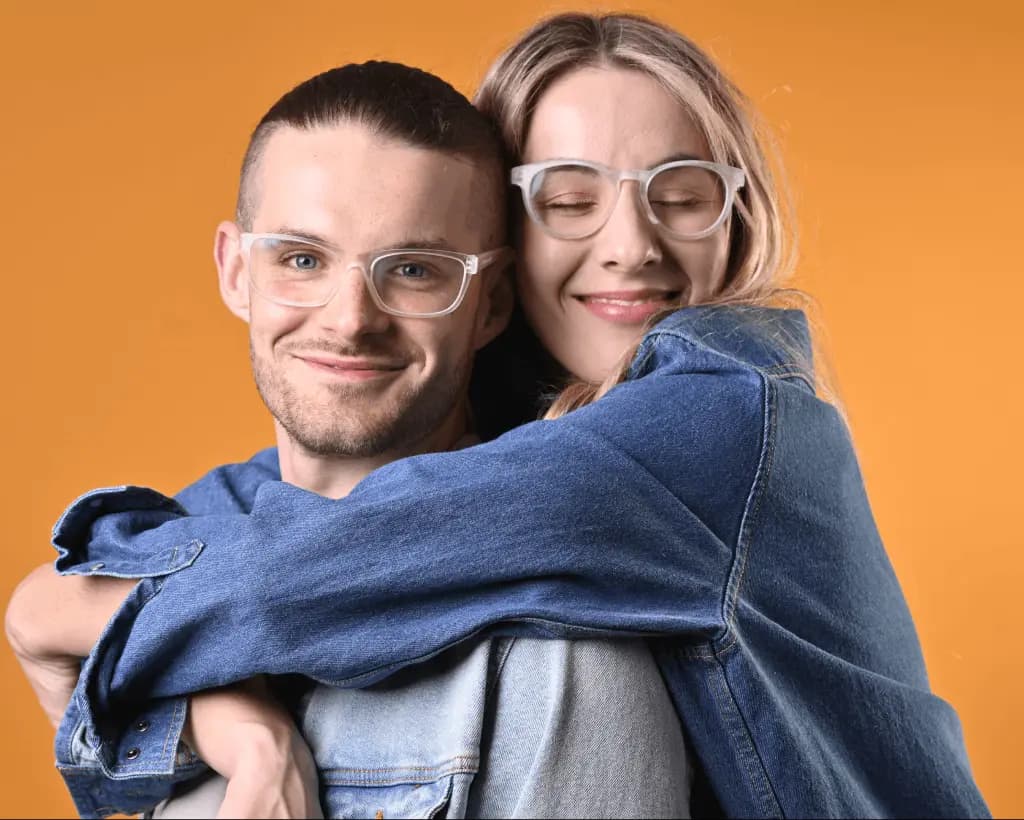 A close-up portrait of two people wearing Dresden Vision glasses, sharing a friendly moment against a warm orange backdrop