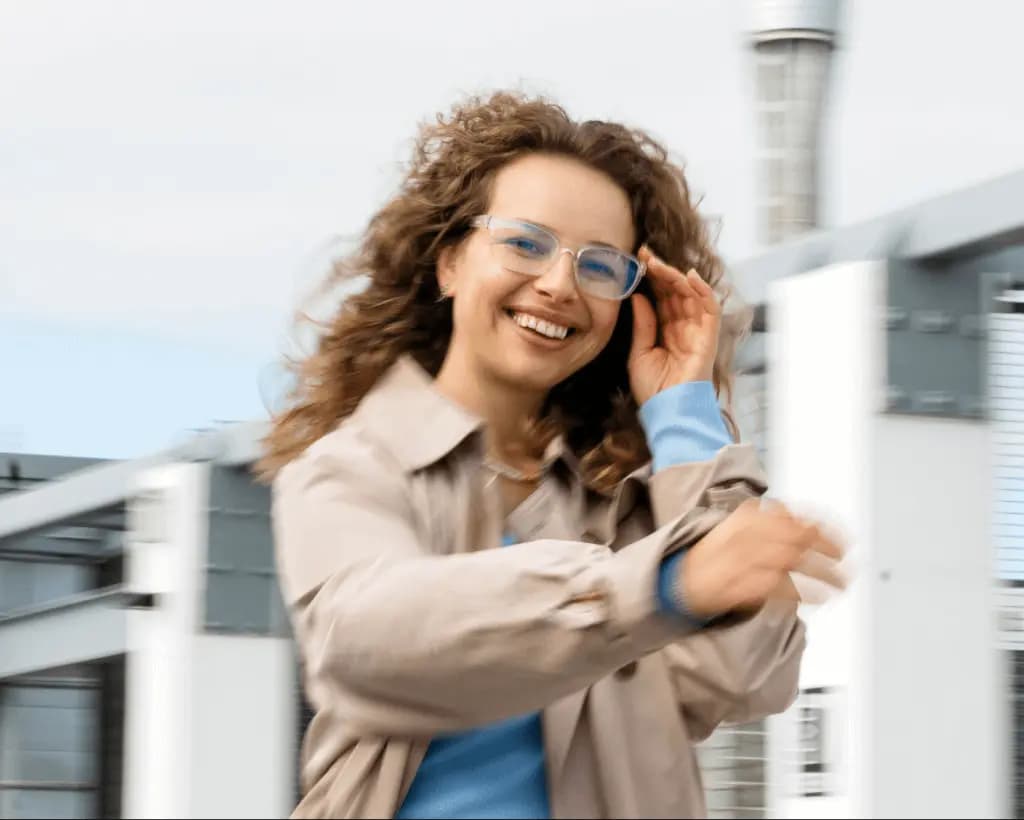 Dynamic portrait of a woman wearing a Dresden Vision glasses and a light coat, smiling while moving outdoors near a modern building