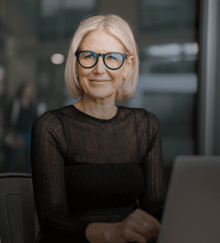 Smiling older woman wearing blue eyeglasses by Dresden Vision Australia, seated indoors with a laptop, representing comfort and clarity with multifocal lenses