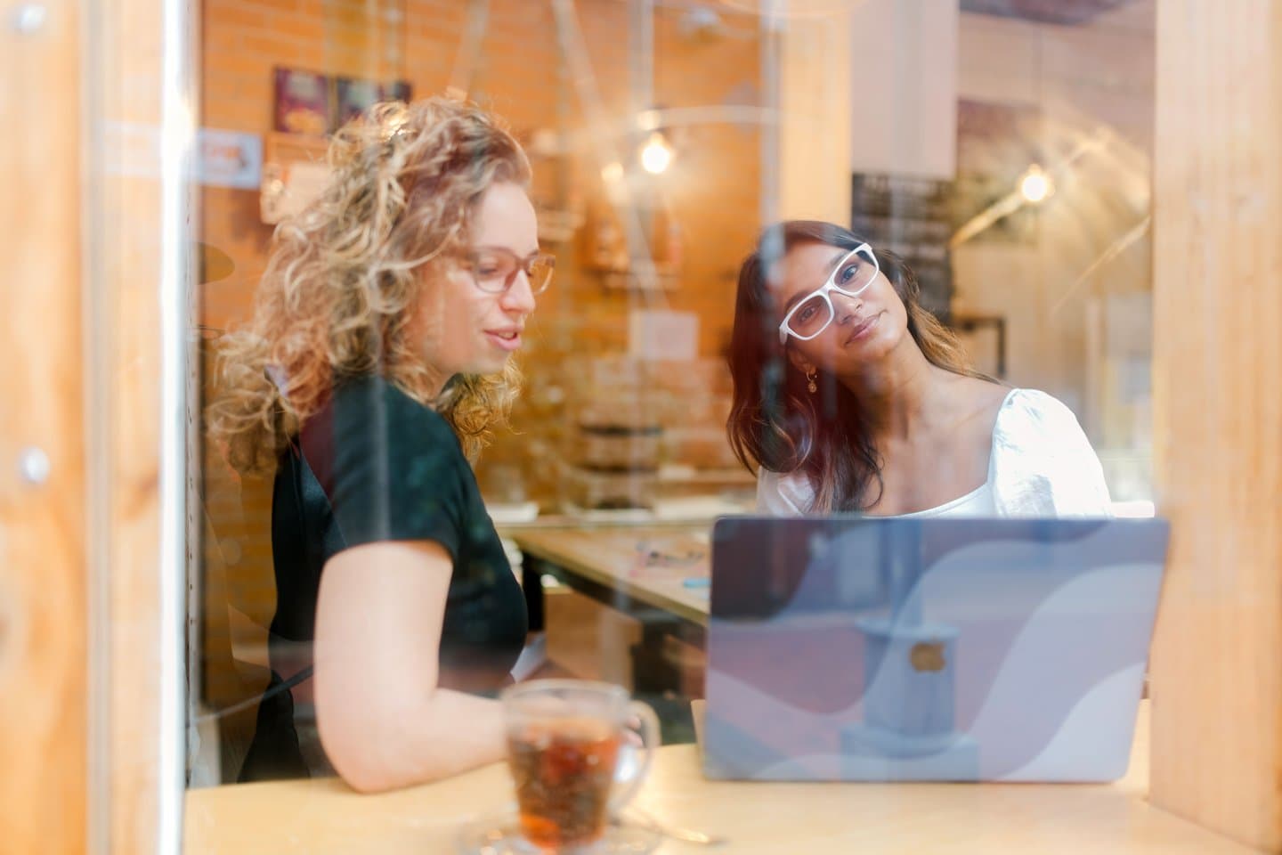 Two women in Dresden Vision Canada glasses enjoy a collaborative moment in a café, with natural light and reflections adding a soft atmosphere.