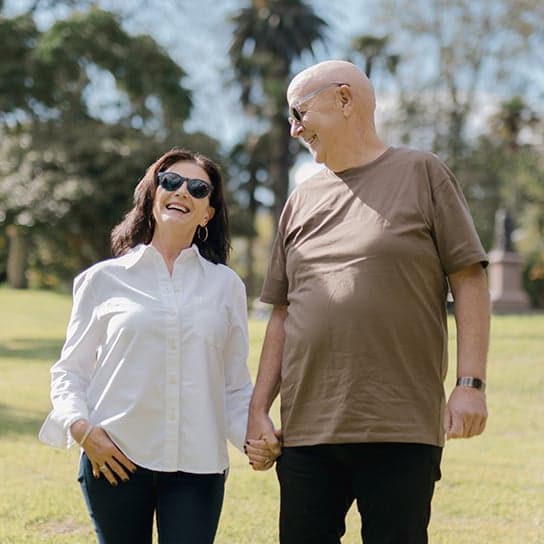 A smiling elderly couple wearing stylish sunglasses holds hands while walking in a sunny park, captured in a joyful and candid moment – Dresden Vision Australia.