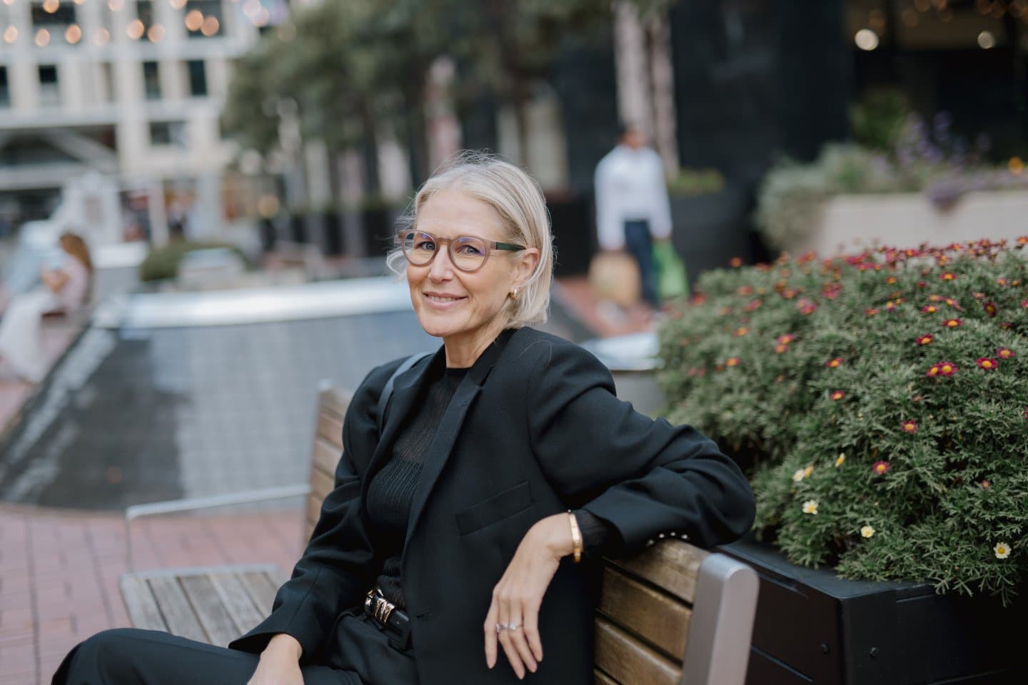 Portrait of a woman wearing sustainable, round-framed glasses by Dresden Vision. She is seated outdoors in a modern urban setting, highlighting the blend of fashion and functional multifocal lens technology.
