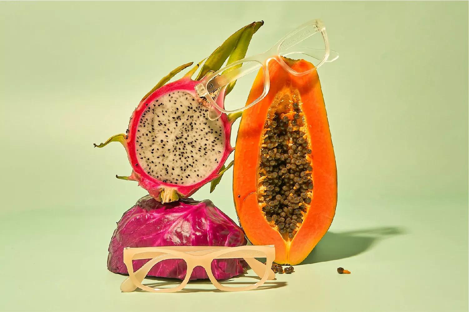 A dragon fruit and papaya sliced in half. A clear Dresden eyeglasses on top of the papaya fruit and dragon fruit, and on the ground a cream colored Dresden eyeglasses.