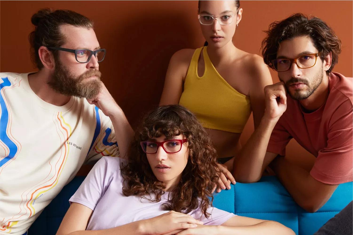 A group of people, two men and two women, all wearing Dresden eyeglasses. The bearded man on the left side is not facing the camera, he has a black Dresden eyeglasses and a vertically striped shirt. The curly semi-long haired man the right facing the camera wearing a red Dresden glasses and a red shirt. The woman on the front with a curly hair wearing a red Dresden eyeglasses wearing a purple shirt. A woman at the back wearing a yellow top and a clear Dresden eyeglasses. 