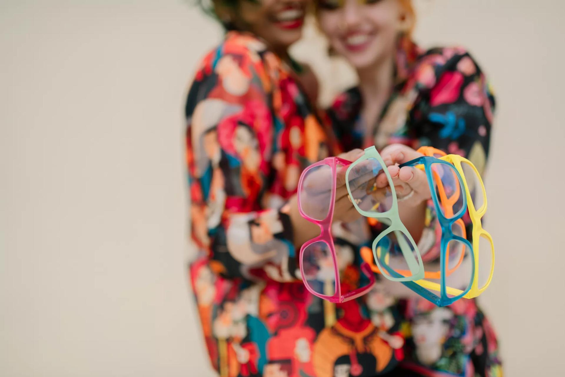 Two individuals in floral print clothing displaying Dresden Vision eyeglass frames in pink, mint green, blue, and yellow. Highlighting the bright frames from Dresden Vision Canada.