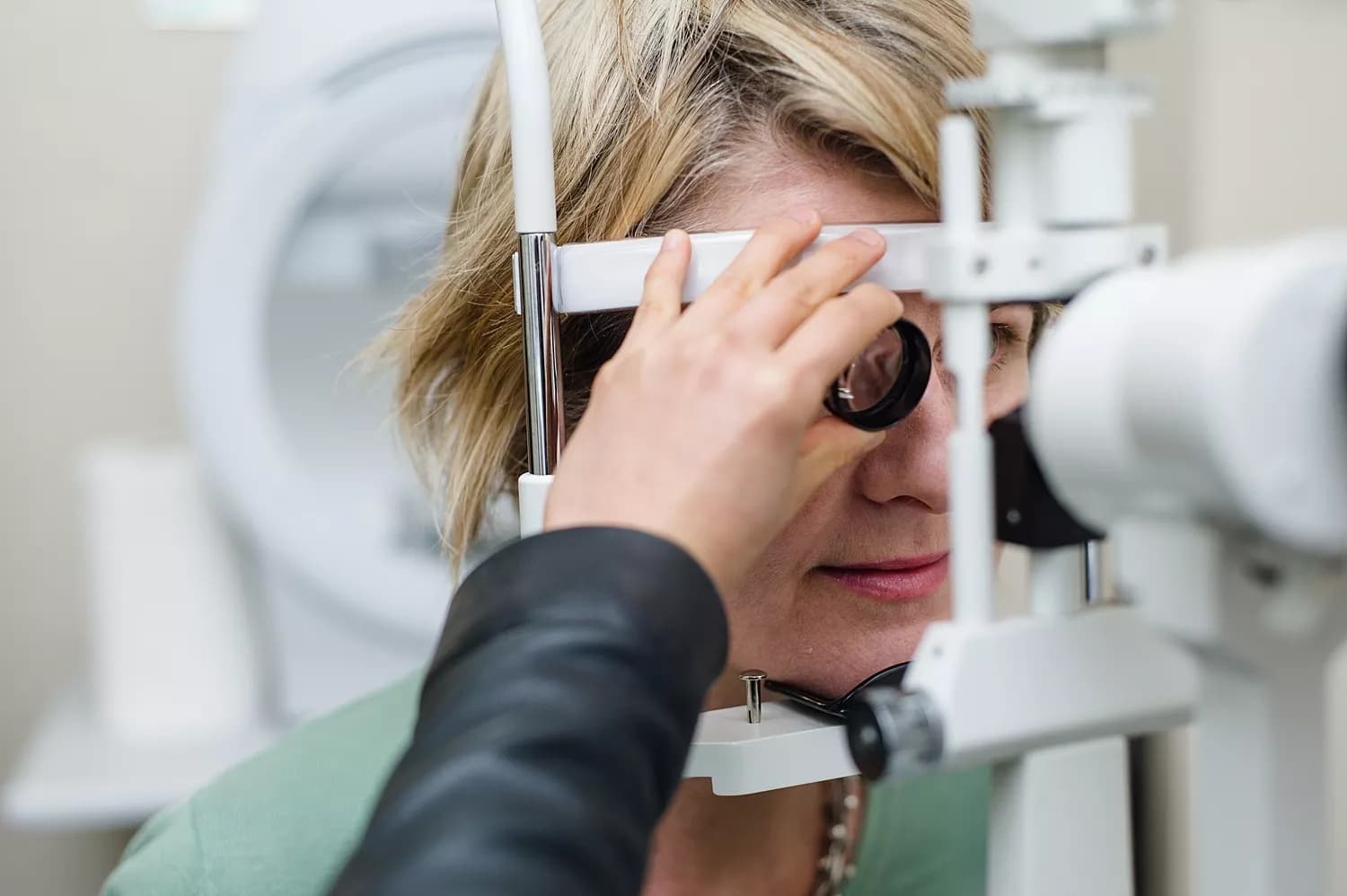 A detailed view of an eye examination at a Dresden Vision clinic in New Zealand. A blonde-haired patient is focused on an optometric device as the optometrist adjusts the equipment. The patients eye can be seen through the lens, with blurred medical tools in the background, highlighting the professional environment.