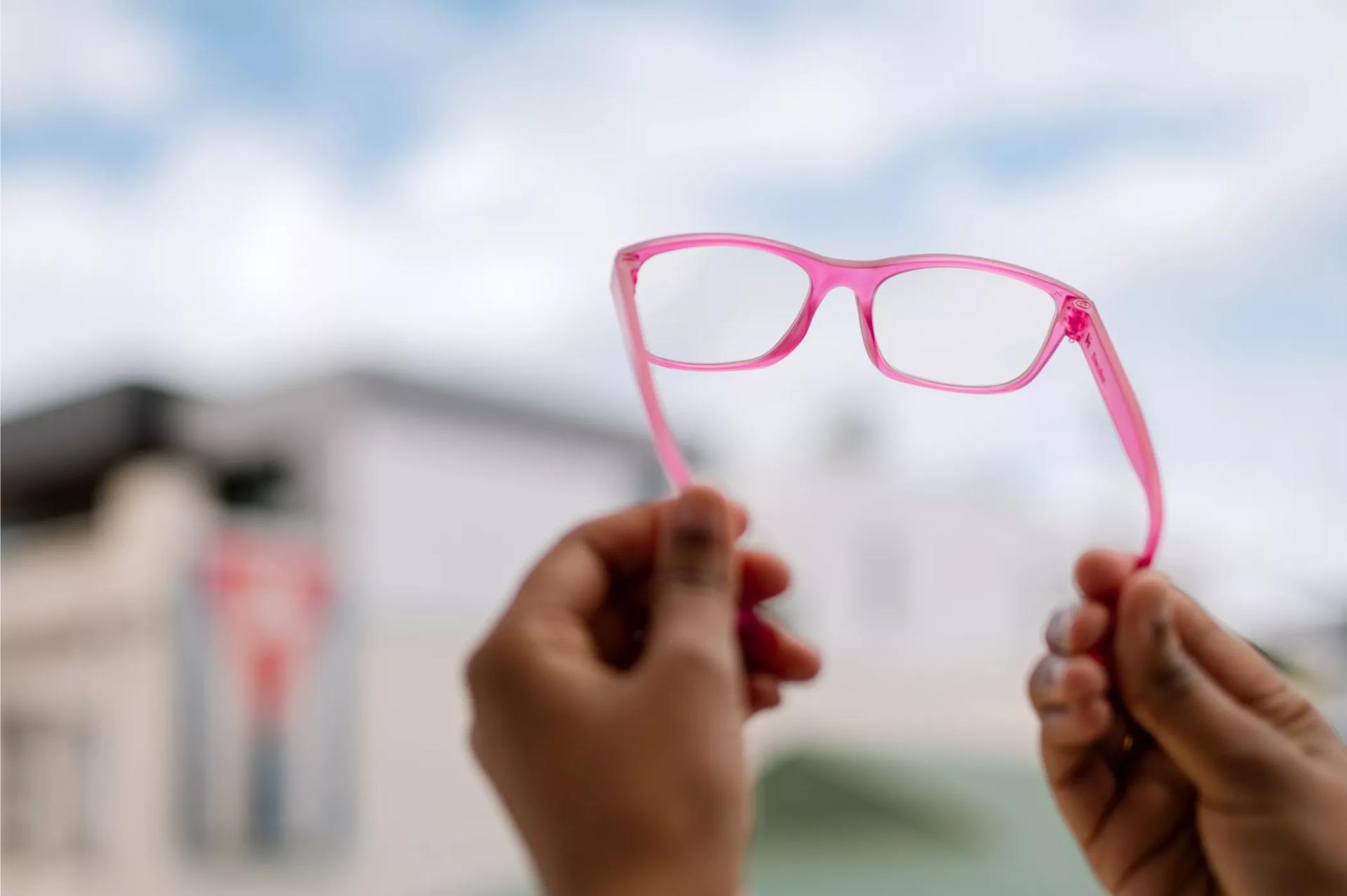 Pink-framed Dresden glasses held up by hands, viewed against a blurry urban background and cloudy sky.