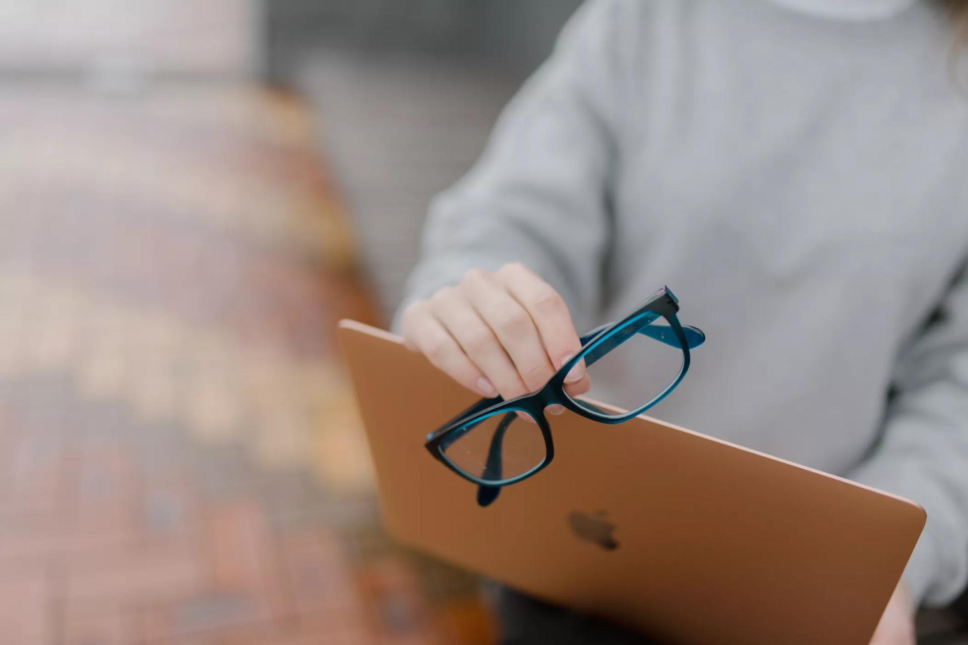 Close-up of hands holding blue rectangular eyeglasses from Dresden Vision New Zealand over a rose gold laptop, demonstrating computer glasses for screen use.