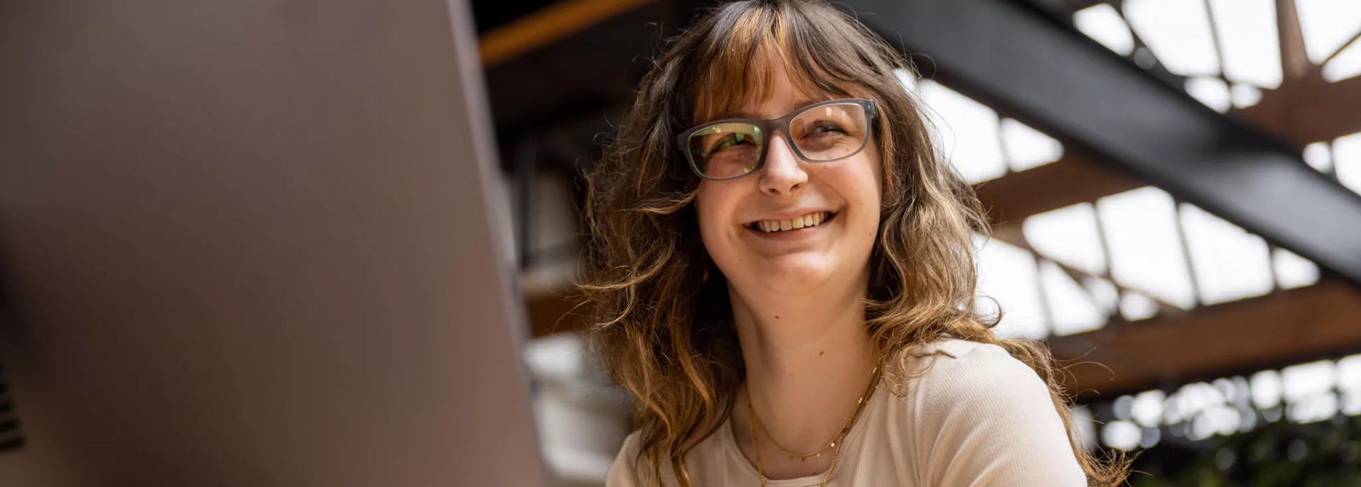 A woman with wavy hair and bangs sits indoors, smiling warmly while wearing stylish multifocal lenses.