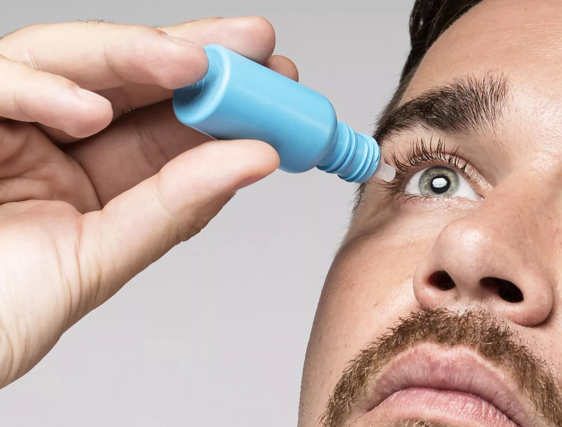 Close-up of a person applying eye drops to their right eye, showing part of their face with a mustache. The hand holds a light blue eye drop bottle, positioned above the open eye, with Dresden Vision Australia eyewear protection in mind.