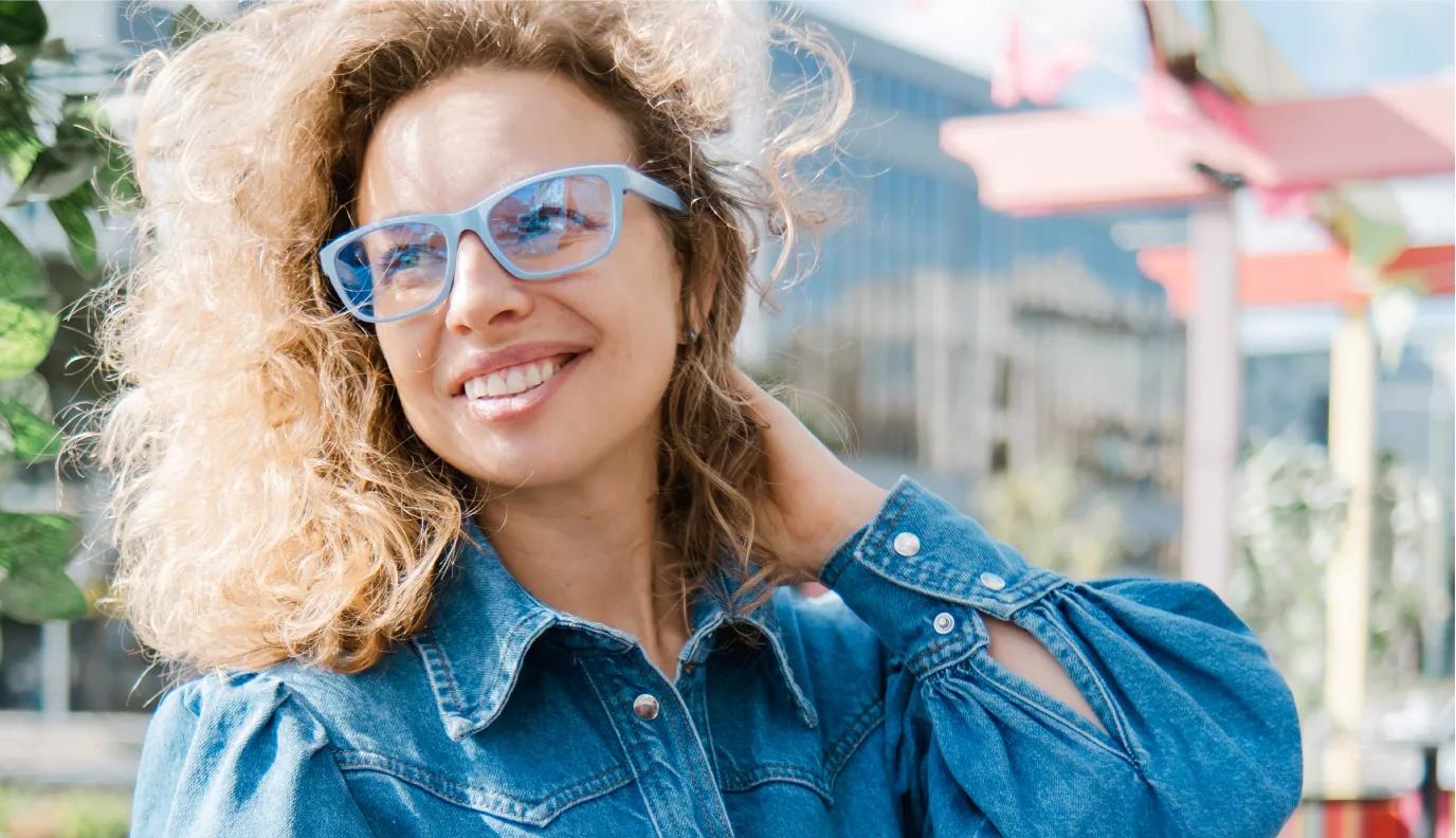 Smiling blonde woman with curly hair wearing blue Dresden glasses and denim jacket, photographed outdoors in urban setting.