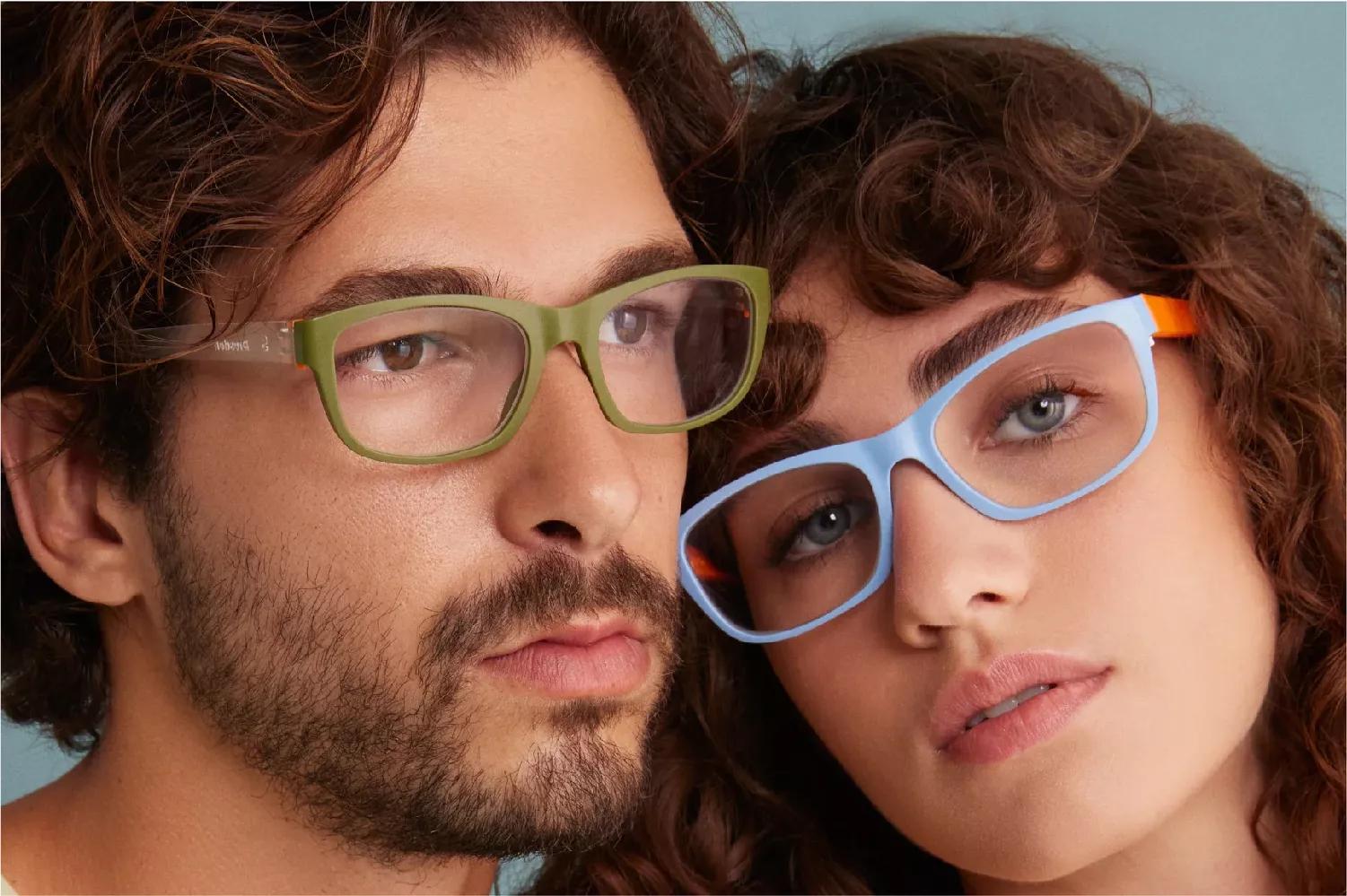 A man and a woman wearing Dresden eyeglasses, the man with a beard and curly hair wears green framed with clear armed Dresden eyeglasses , and the woman with curly hair wears blue framed and orange armed Dresden eyeglasses.