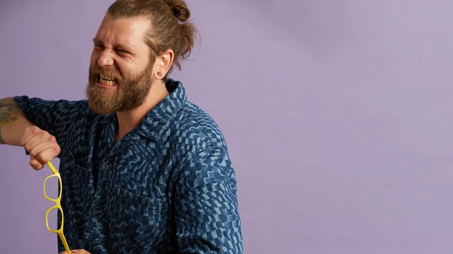 A man with a messy top knot hairstyle wears a blue patterned shirt while proudly displaying bold yellow-framed Dresden Vision glasses in Canada. The light purple background enhances the appeal of the glasses, showcasing their unique design.