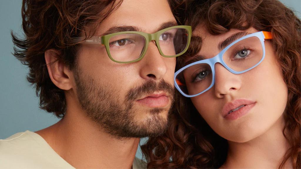 A man and a woman wearing Dresden eyeglasses. The man have a beard and a mustache, he wears a green framed Dresden eyeglasses, and the woman with a curly hair wears a blue Dresden eyeglasses.