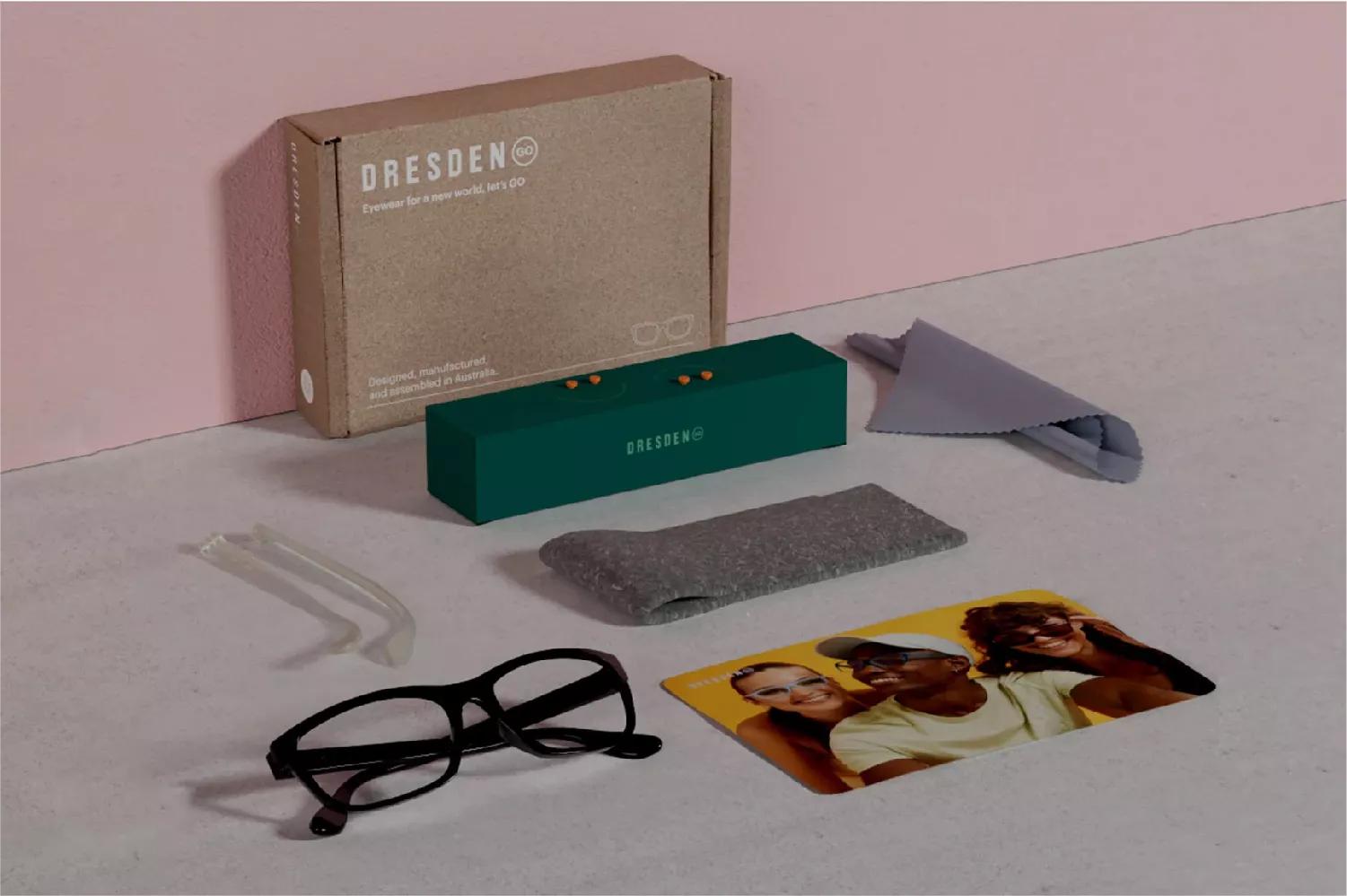 A Dresden kit that contains a black Dresden eyeglasses, an extra pair of arms that is have a clear color, wipes, 2 sets of color orange pins placed above a rectangular green box, a felt case, and photocard having three people smiling, 2 women on the side, and a man in between. 