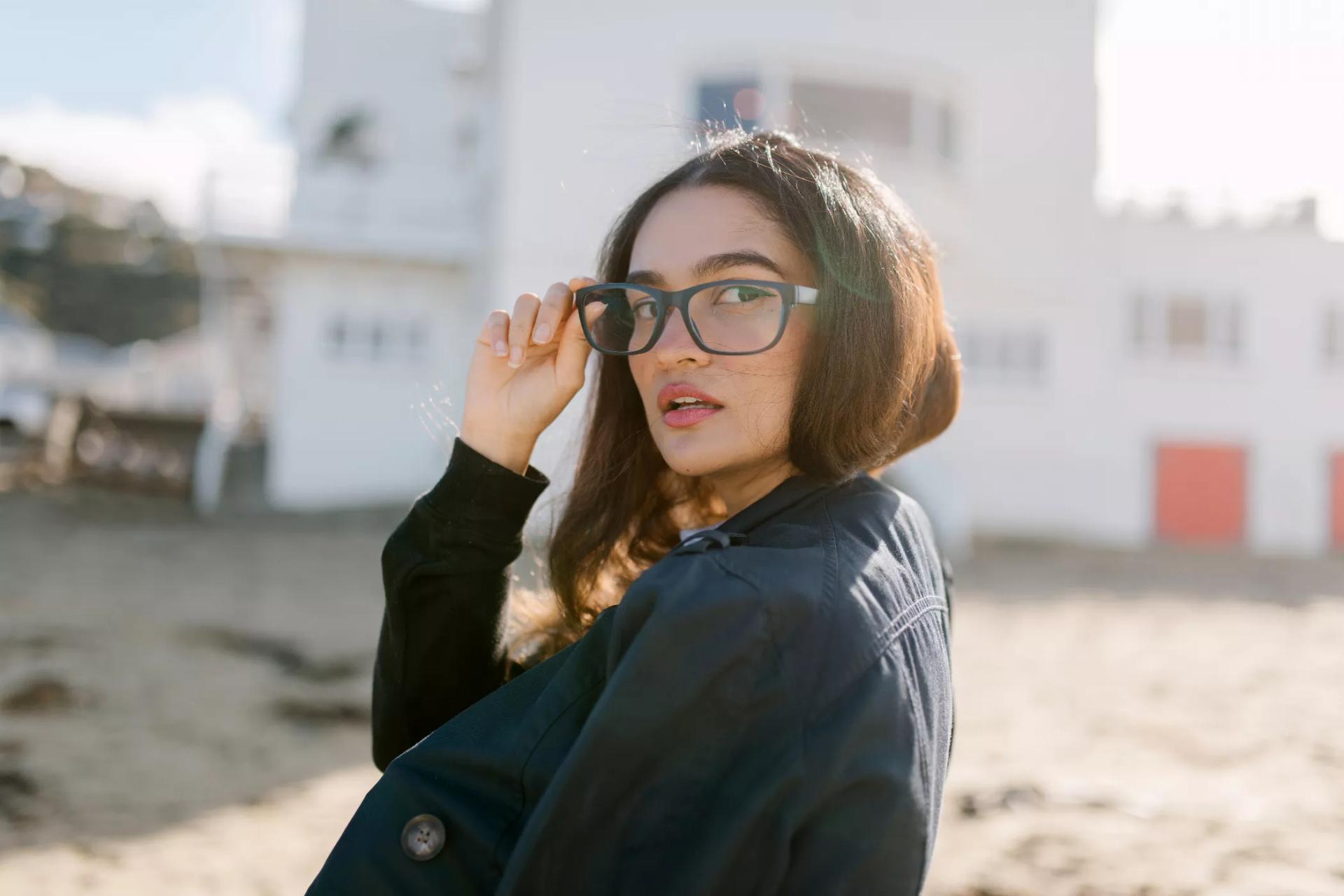 A woman wearing black jacket and a black framed Dresden eyeglasses posing for a picture outdoors under the sun with blurry building in the background