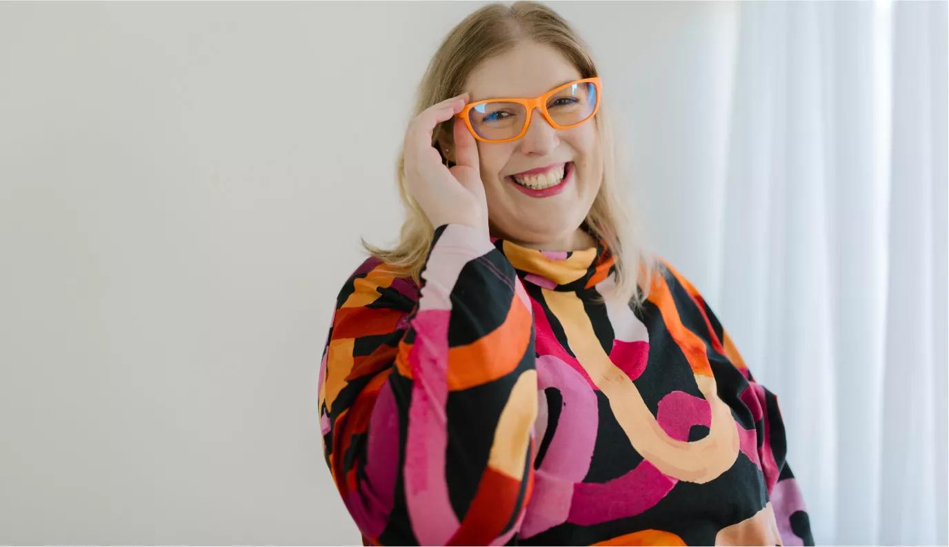 Smiling blonde woman with wearing orange Dresden glasses and a colorful patterned top, photographed indoors near a curtain.