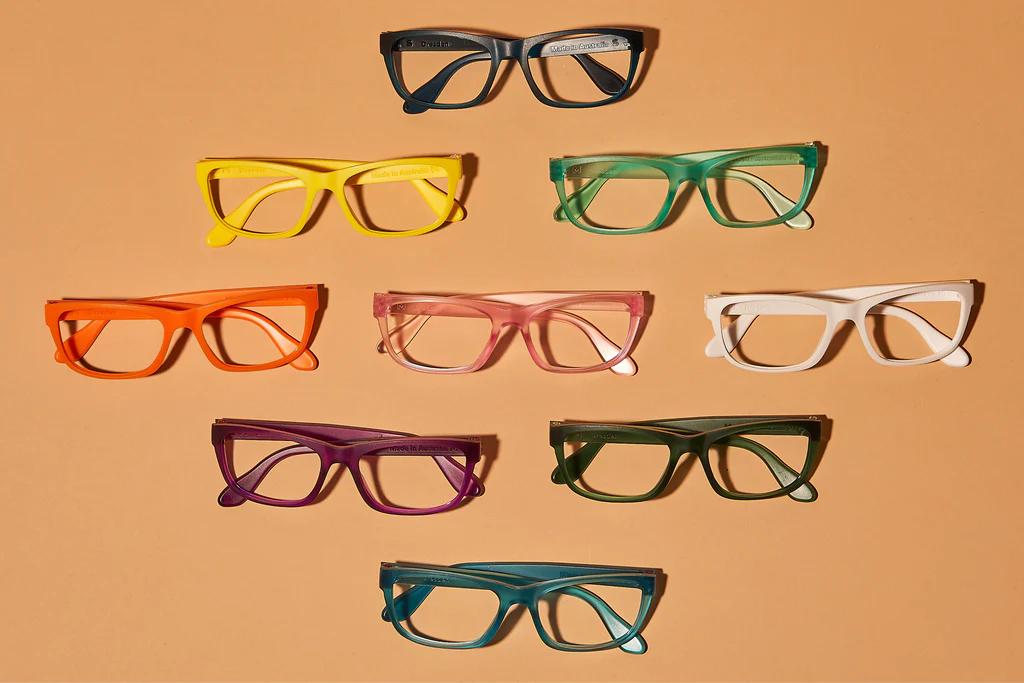 A set of eyeglasses in a diagonal position. There are a total of nine eyeglasses in a variety of colors which are black, yellow, green, orange, pink, cream, violeet, dark green, and blue.