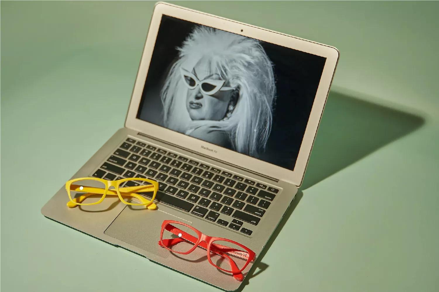 A brown Macbook with a pair of Dresden glasses on top of it. There are two Dresden eyeglasses, one being color red, and the other one is yellow. In the screen of the Macbook is black and white picture of an old woman wearing white eyeglasses with black lenses. Her hair is stylish but messy. 