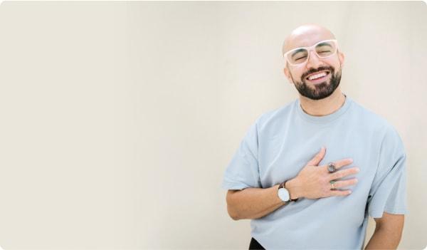 A happy, bearded man with a shaved head wears Dresden Vision glasses and a light blue shirt. His warm smile and hand-to-chest pose convey the pride and comfort found in choosing eyewear in Canada.