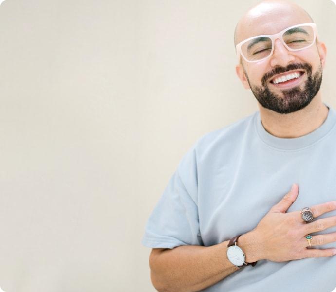 A happy, bearded man with a shaved head wears Dresden Vision glasses and a light blue shirt. His warm smile and hand-to-chest pose convey the pride and comfort found in choosing eyewear in Canada.