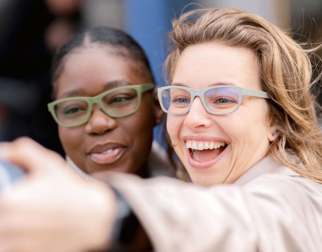 Two friends, both wearing prescription glasses, are taking a selfie together. The woman on the right smiles broadly, showing her teeth, while the woman on the left has a gentle smile. Both appear to be enjoying a happy moment outdoors.