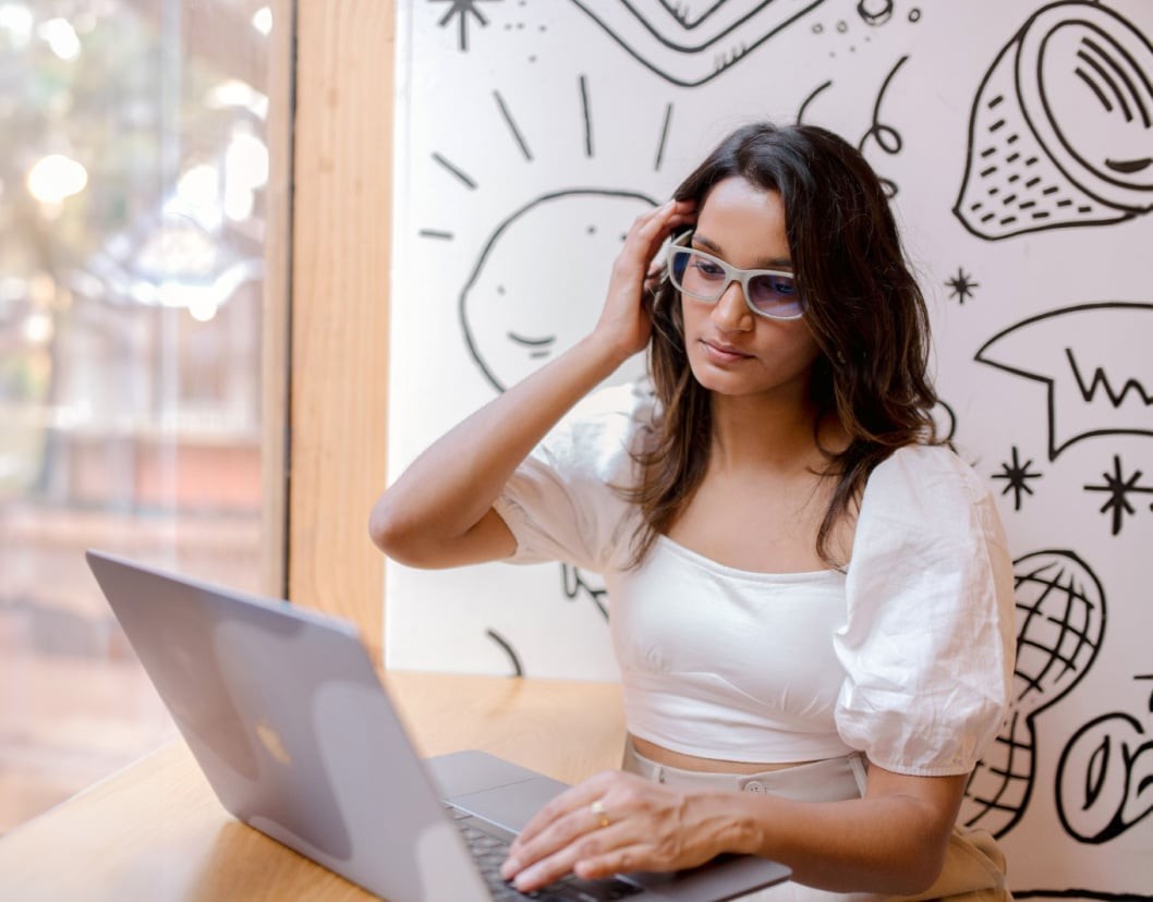 A woman wearing stylish reading glasses in Canada, sitting by a window, working on her laptop with artistic wall designs.