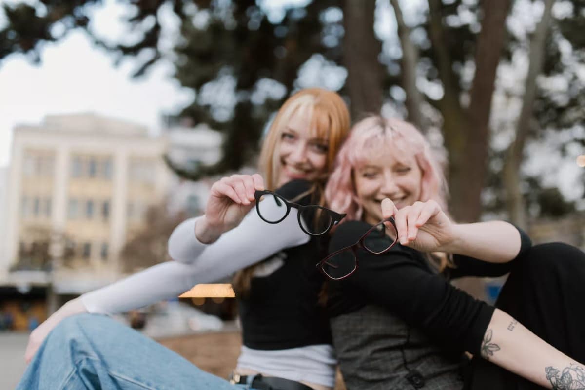 Smiling duo showing off their colourful frames with Single Vision Lenses from Dresden Vision New Zealand, enjoying a laid-back day beneath city trees.