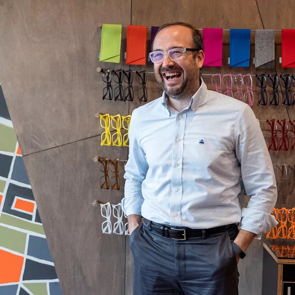 A smiling bearded man wearing glasses stands in a Dresden Vision eyewear store. Behind him, colorful Dresden frames are displayed on the wall, alongside vibrant fabric swatches. The store's interior showcases Dresden's modern and stylish approach to eyewear retail.
