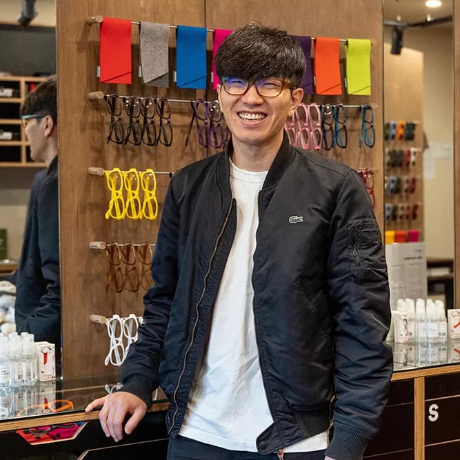 Smiling young man wearing glasses and a dark jacket in an eyewear store, with colorful Dresden frames displayed on the wall behind him.