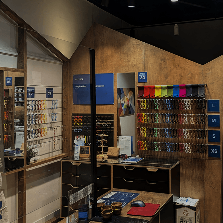 Wood-paneled interior of Dresden Vision Riverside in Toronto. A green door features the logo and "Glasses that don't cost the earth." Colorful frames are neatly displayed by size, marking the closest optometry clinic to its neighbor locations.